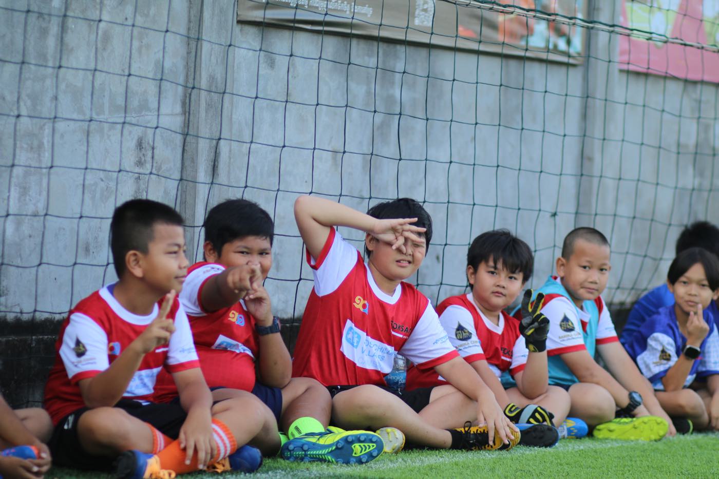 A row of young players in red kit sitting against the goal net, waiting their turn